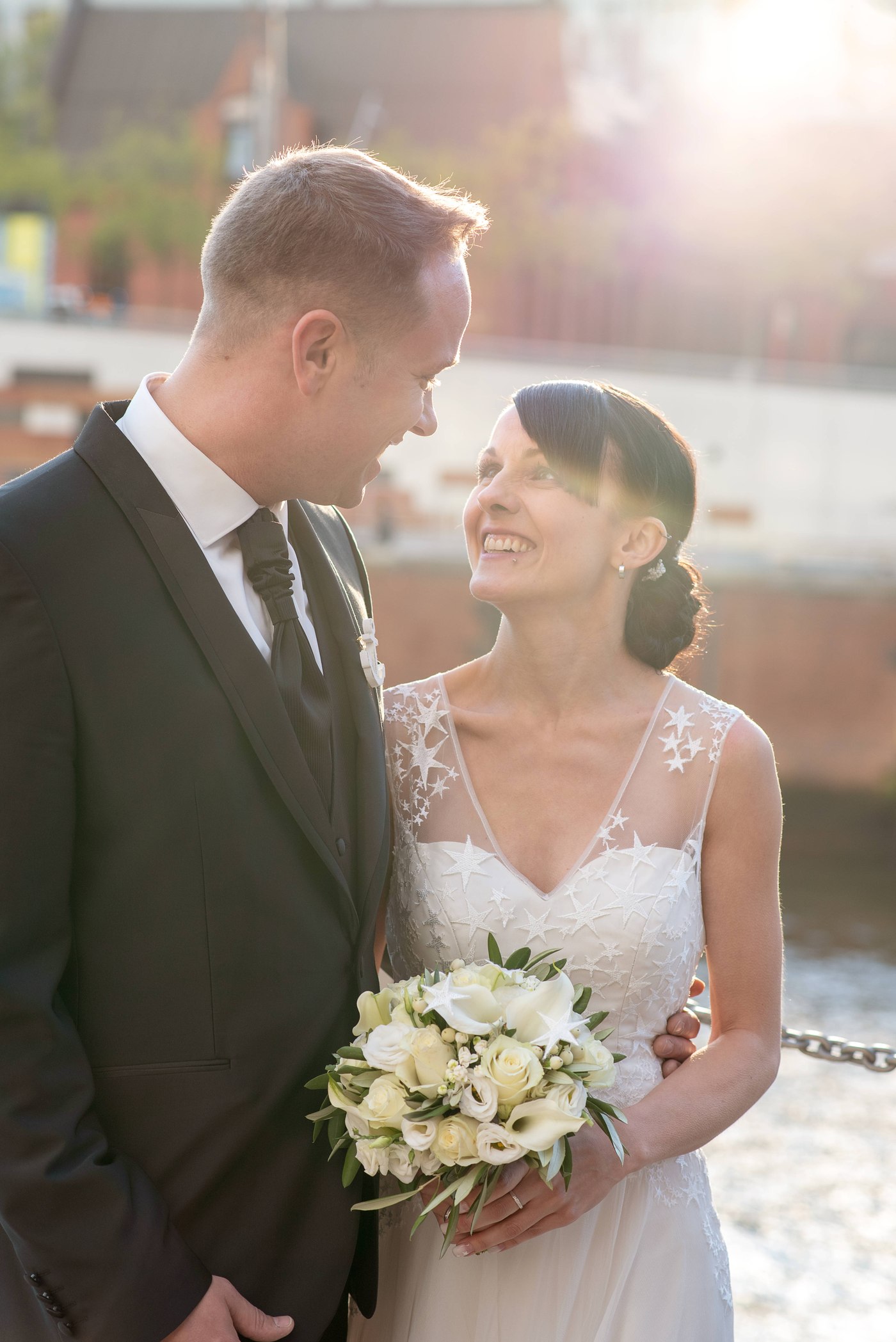 John Eid und seine Frau Tini lächeln sich am Hochzeitstag an, sie hält den Brautstrauß, Gegenlicht am Hafen